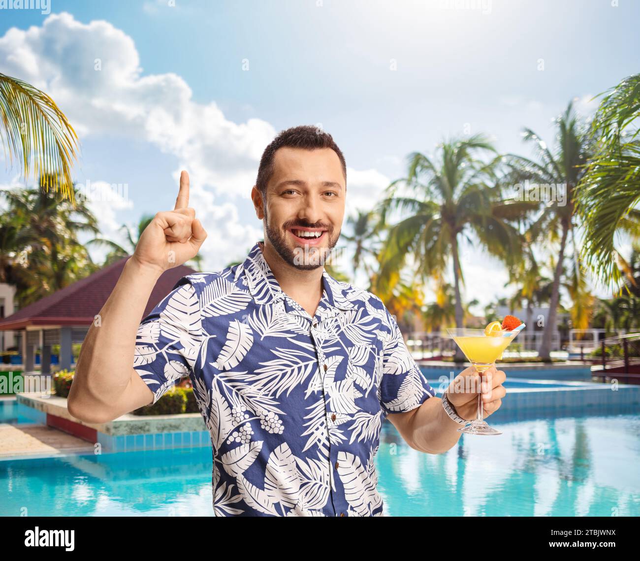 Junger Mann, der ein Glas Cocktail hält und vor ein Schwimmbad zeigt Stockfoto