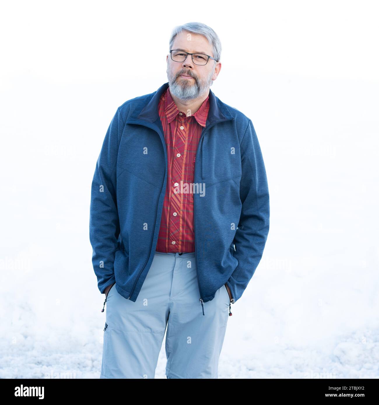 Mann, 56 Jahre, graues Haar und Bart, im Winter draußen mit Schnee im Hintergrund, Hände in der Tasche, Brille tragen Stockfoto