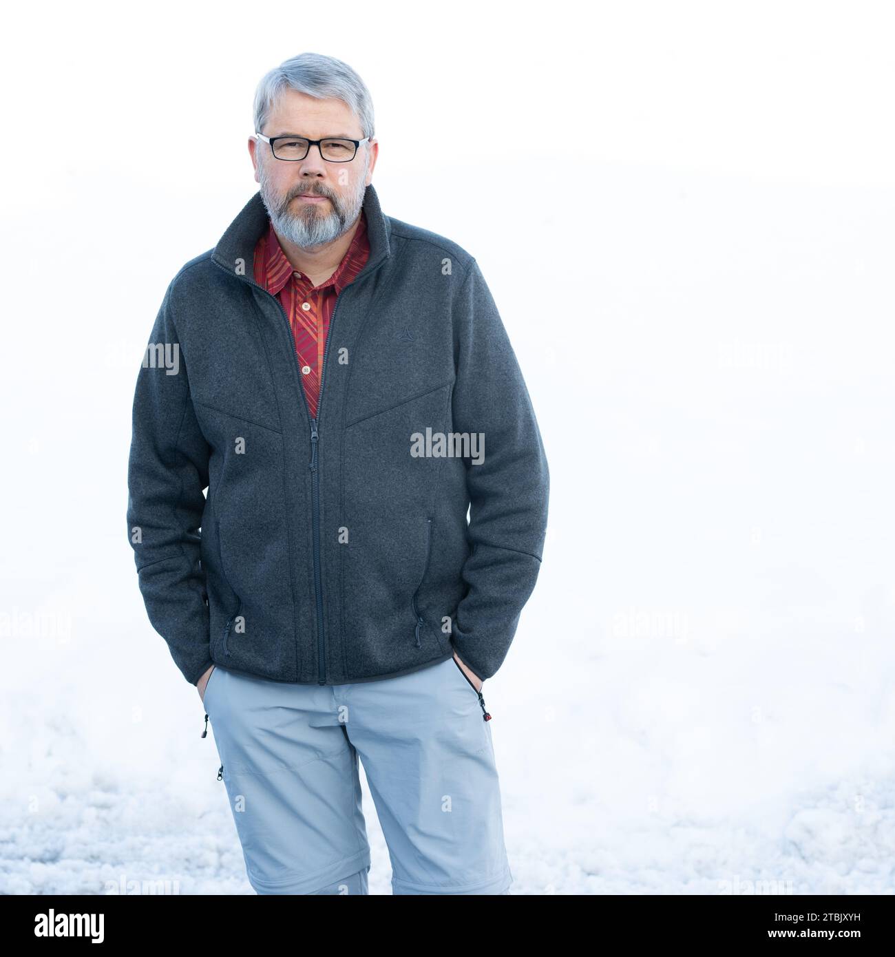 Mann, 56 Jahre, graues Haar und Bart, im Winter draußen mit Schnee im Hintergrund, Hände in der Tasche, Brille tragen Stockfoto