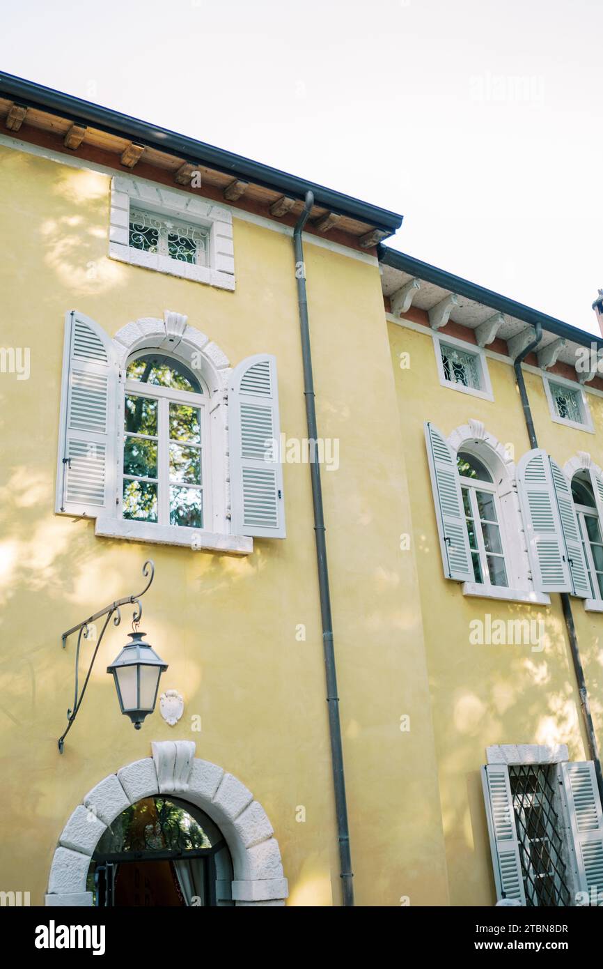 Kleine rechteckige Fenster unter dem Dach an der gelben Fassade eines alten Herrenhauses über Bogenfenstern mit weißen Fensterläden Stockfoto