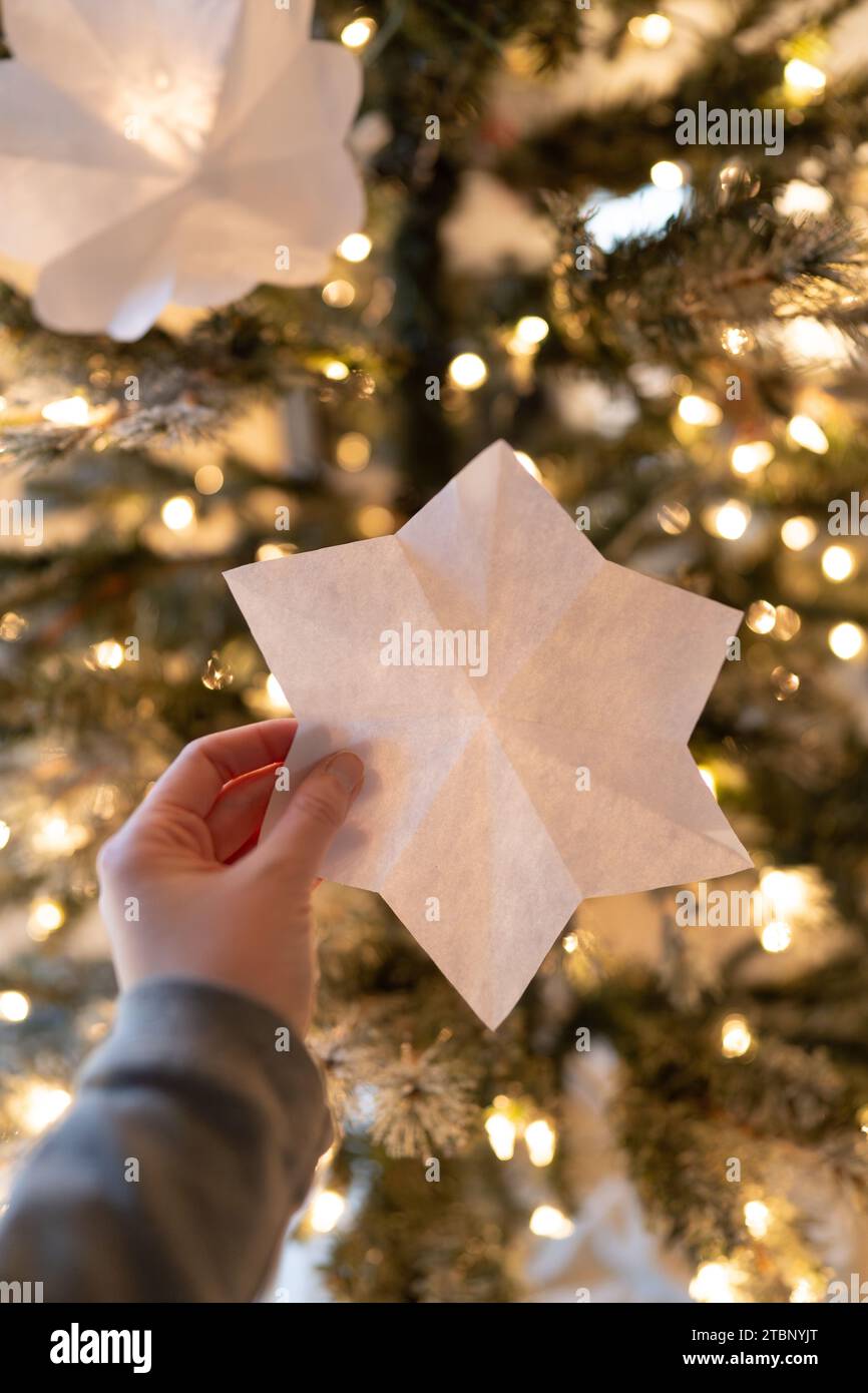 Frau hält handgefertigte Papierschneeflocke vor dem Weihnachtsbaum hoch Stockfoto
