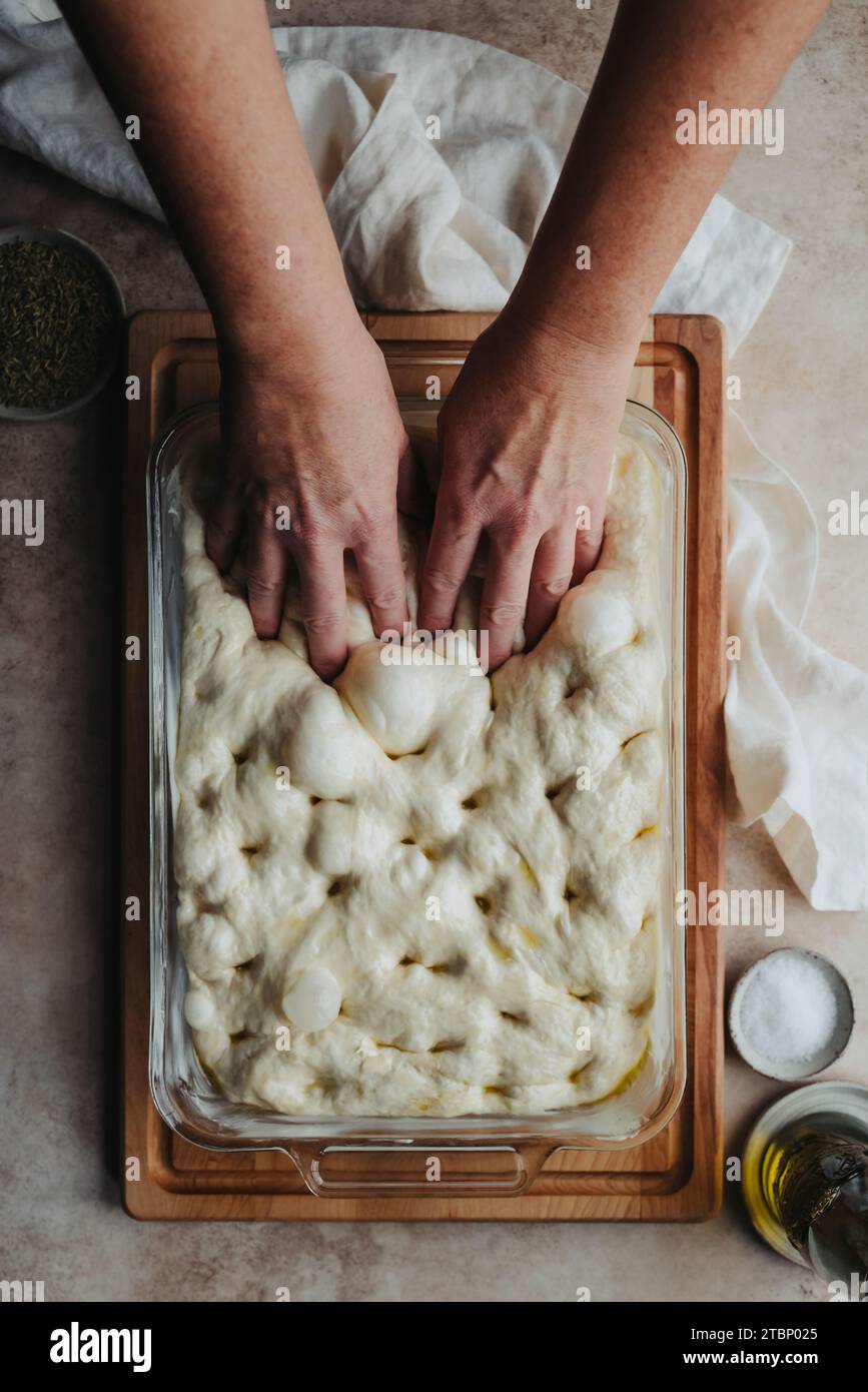 Hände, die Löcher in aufgerissenen Focaccia-Teig machen, von oben geschossen. Stockfoto