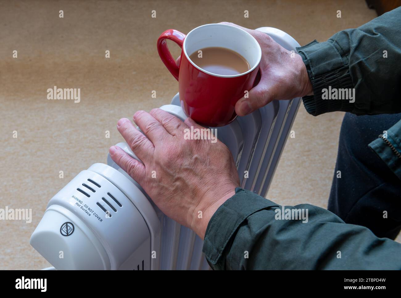 Eine Person, die eine heiße Tasse Tee hält und die Hände auf einem tragbaren, ölgefüllten Heizgerät in einem Wohnzimmer wärmt. Stockfoto