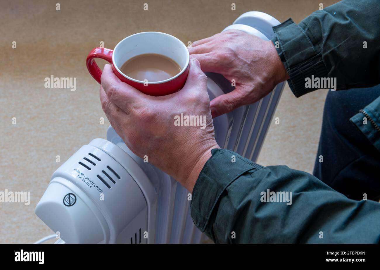 Eine Person, die eine heiße Tasse Tee hält und die Hände auf einem tragbaren, ölgefüllten Heizgerät in einem Wohnzimmer wärmt. Stockfoto