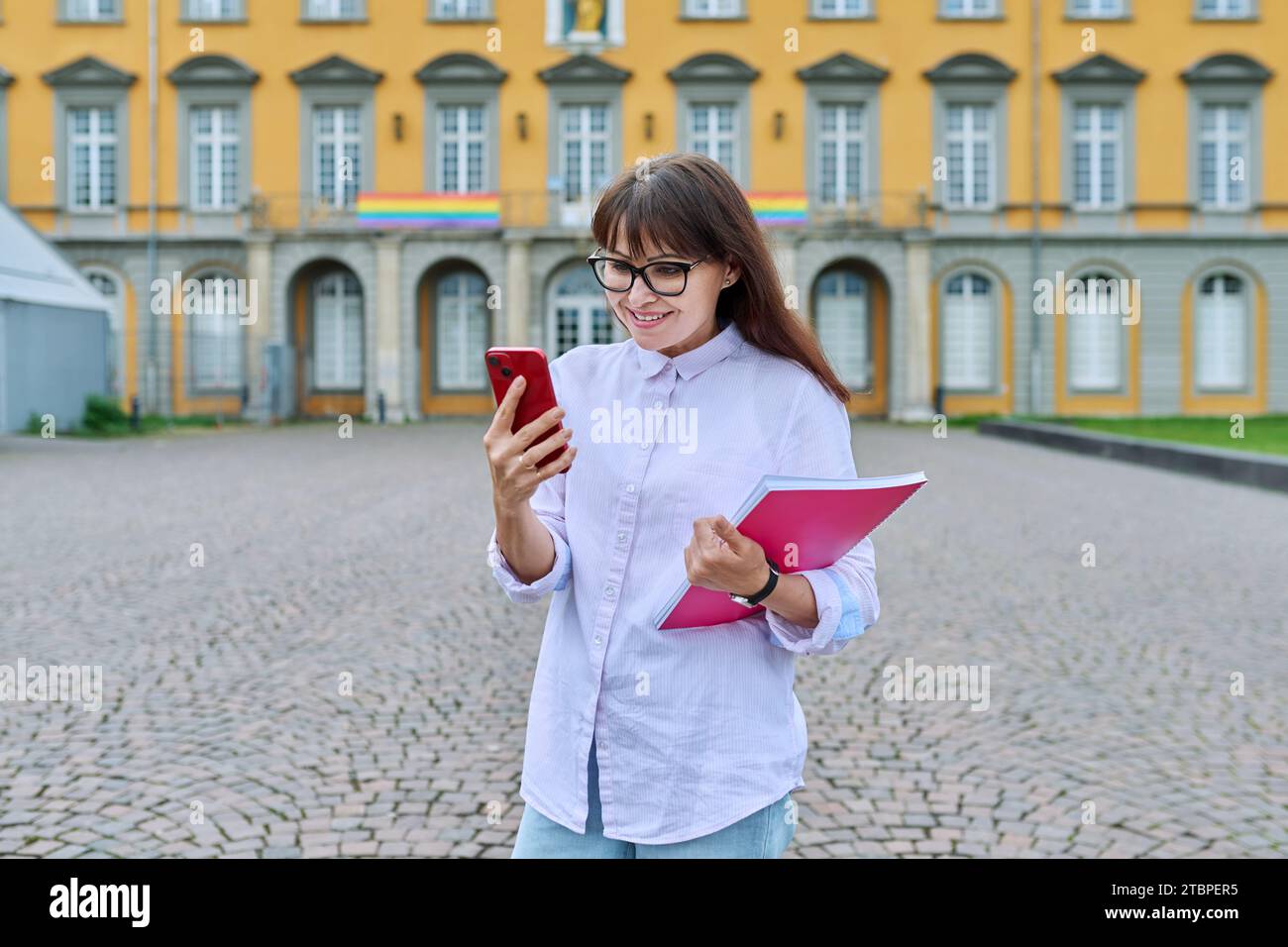 Lehrerin mittleren Alters mit Smartphone, Hintergrund des Bildungsgebäudes Stockfoto