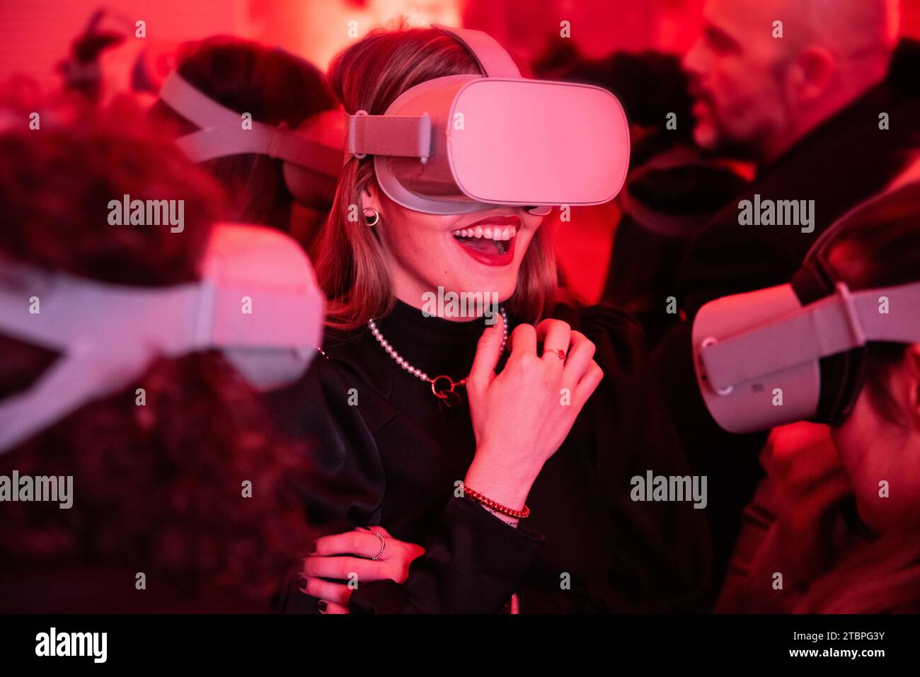 Inmitten eines Meeres von rot beleuchteten VR-Enthusiasten lacht eine fröhliche Frau und verbindet digitale und physische in einem immersiven Erlebnis Stockfoto
