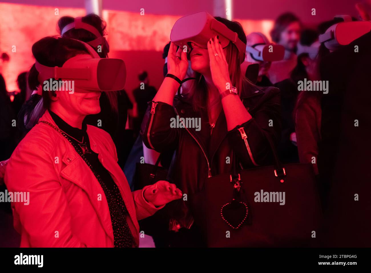Zwei Frauen, die in virtuelle Welten eingetaucht sind, umgeben von einem rot beleuchteten Ambiente und einer Menge von Teilnehmern, die die Freude an futuristischer Technologie in einer dynamischen Atmosphäre teilen Stockfoto