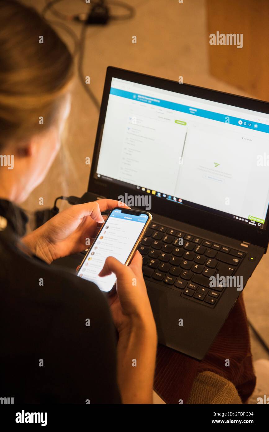 Nahtloses Multitasking: Eine Frau sitzt mit einem Laptop auf dem Schoß, vertieft in eine mobile Chat-App. Eine Momentaufnahme moderner Konnektivität und effizienter Arbeit Stockfoto