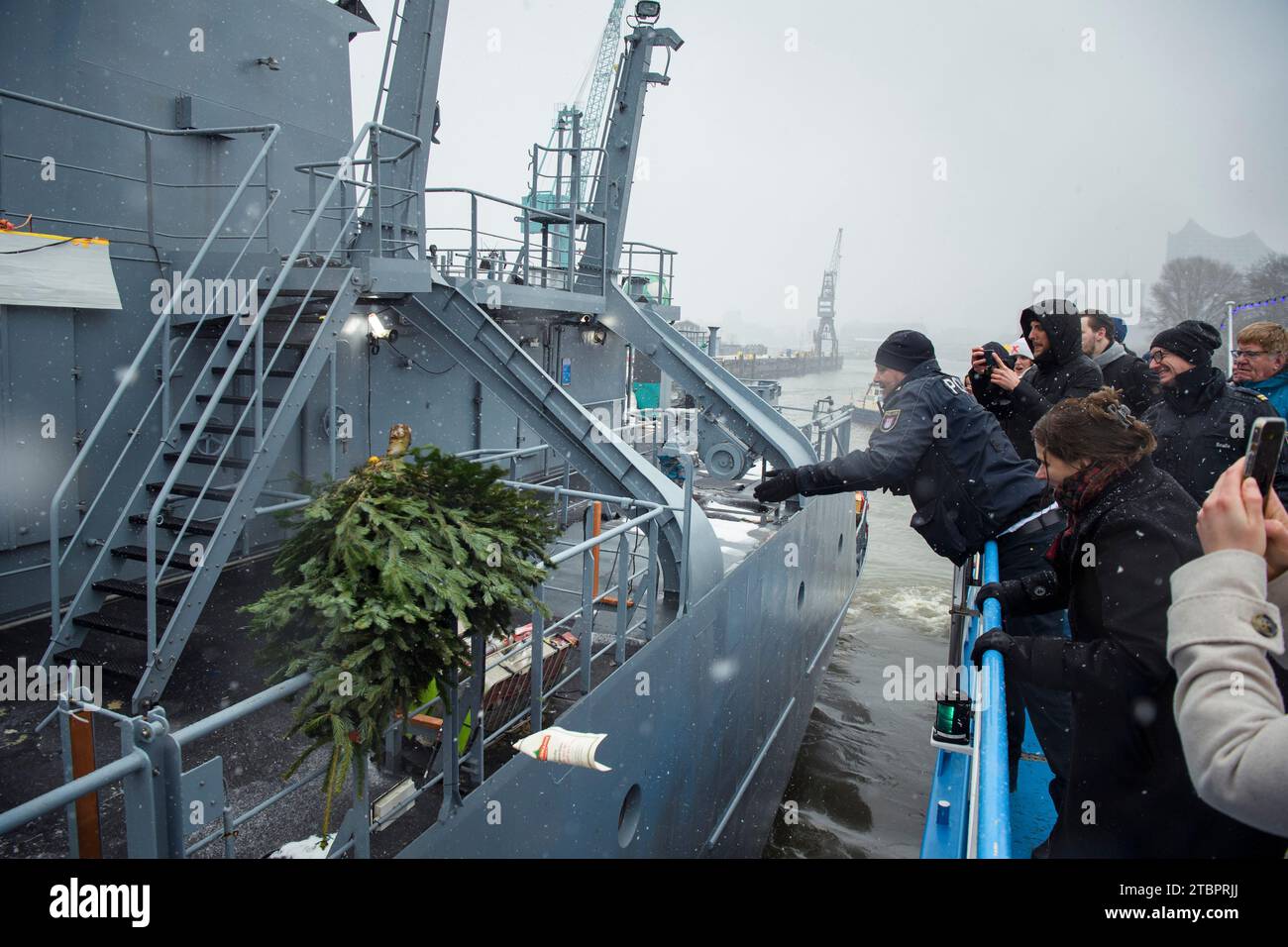 Hamburg, Deutschland. Dezember 2023. Während der Kampagne „Weihnachtsbäume für Schiffe im Hamburger Hafen“, die von der Nordmann-Informationszentrale gemeinsam mit der Hansestadt Hamburg organisiert wurde, wird vom Deck eines Ausflugsschiffes Rainer Abicht ein Weihnachtsbaum auf ein deutsches Marineschiff geworfen. Jedes Jahr Anfang Dezember werden Weihnachtsbäume an Seeleute auf Schiffen im gesamten Hafengebiet geliefert. Quelle: Gregor Fischer/dpa/Alamy Live News Stockfoto