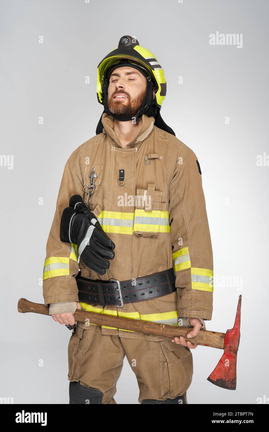 Erschöpfter kaukasischer Feuerwehrmann mit geschlossenen Augen, schäbige Axt in der Hand. Vorderansicht eines müden, bärtigen Mannes in Uniform mit Beil auf grauem Atelierhintergrund. Konzept der Arbeit, des Arbeitsplatzes, der Ausrüstung. Stockfoto