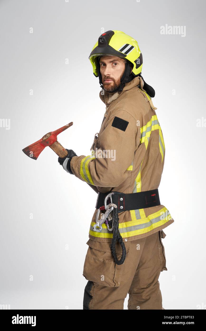 Ernsthafter kaukasischer Feuerwehrmann mit Helm und großer Axt im Studio. Blick von der Schulter auf einen bärtigen männlichen Retter mit Beile, Blick in die Kamera, auf grauem Hintergrund. Arbeitskonzept, Feuerwehrausrüstung. Stockfoto