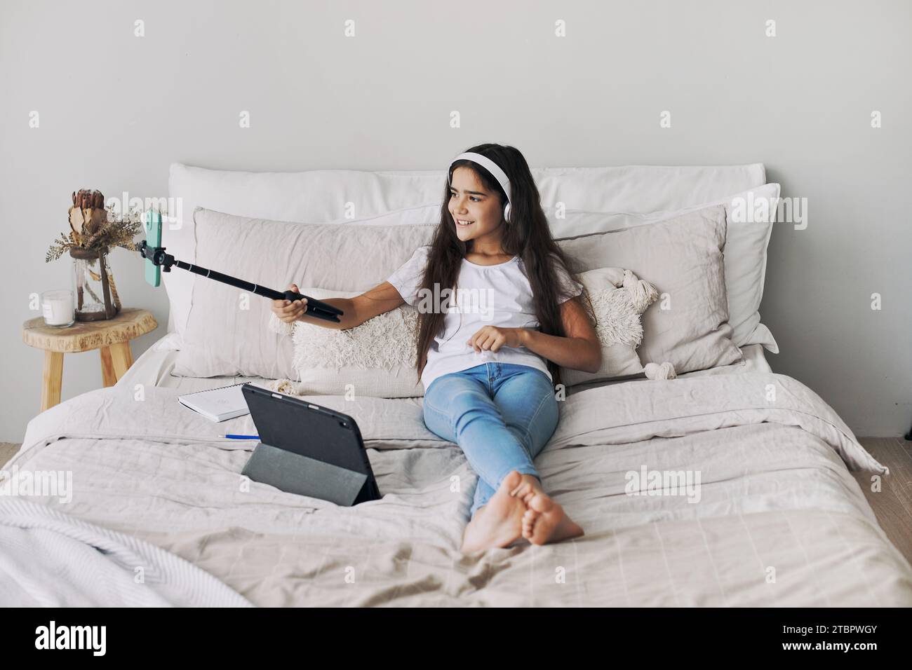 Attraktives Mädchen, das auf dem Bett im Schlafzimmer sitzt und Selfie-Stick hält, Fotos machen, Videos für Social-Media-Kanäle aufnehmen, an Live-s teilnehmen Stockfoto