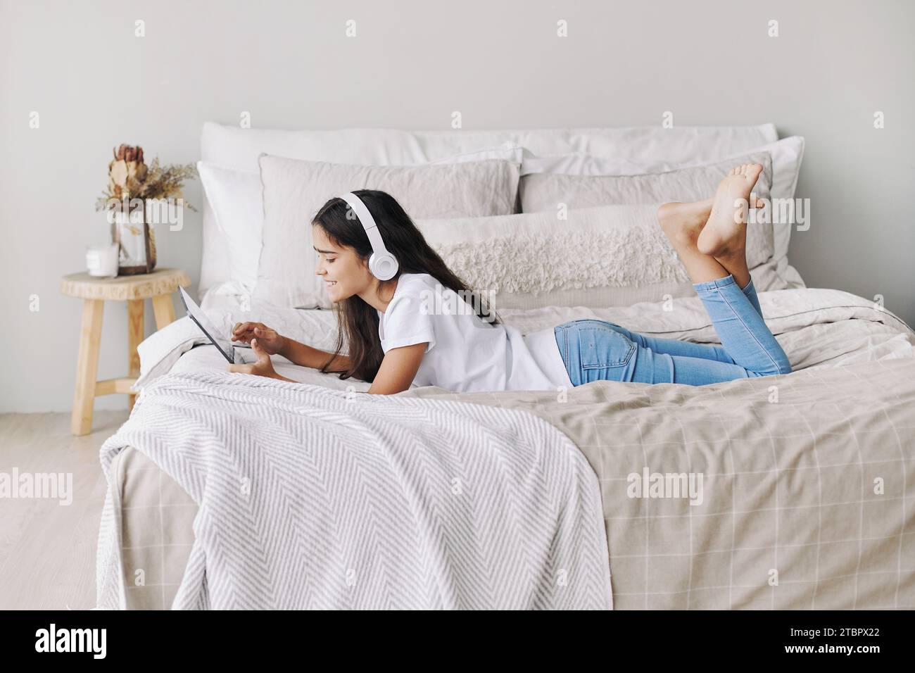 Entzückendes Mädchen aus Jugendzeit in kabellosen Kopfhörern, das mit einem digitalen Tablet auf dem Bett liegt. Junge Generation und moderne Technik überbeanspruchen, Spielgeräte für Spaß, Stockfoto