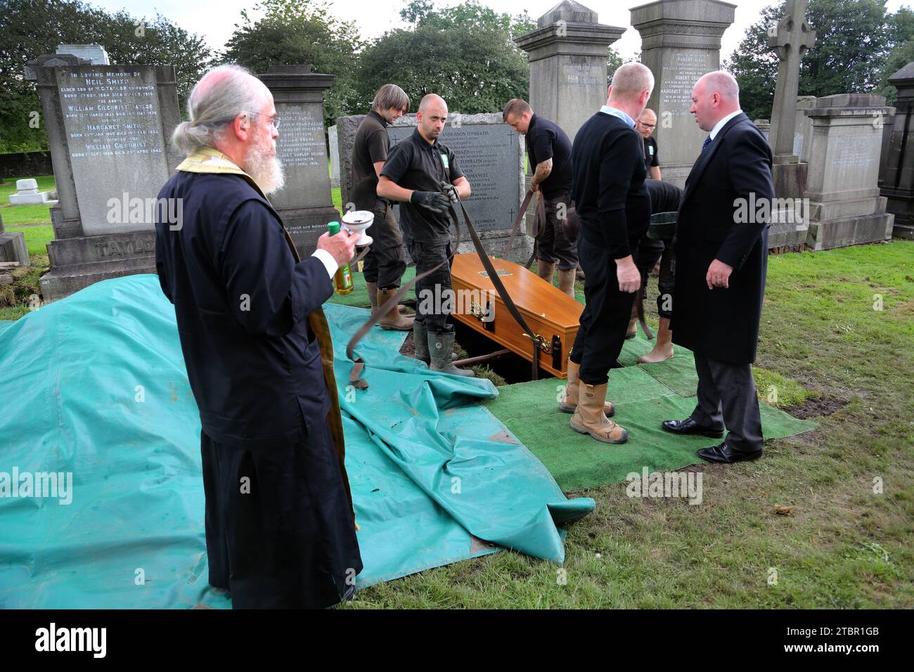 Glasgow Schottland Griechisch-orthodoxe Beerdigung in Glasgow Necropolis - Priest hält eine Flasche Olivenöl, während der Sarg manuell in das Grab gesenkt wird Stockfoto