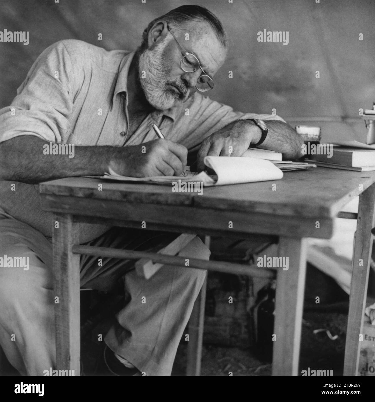 Foto von Ernest Hemingway, der um 1953 auf seinem Campingplatz in Kenia an einem Tisch sitzt und schreibt. Publisher Look Magazine. Stockfoto