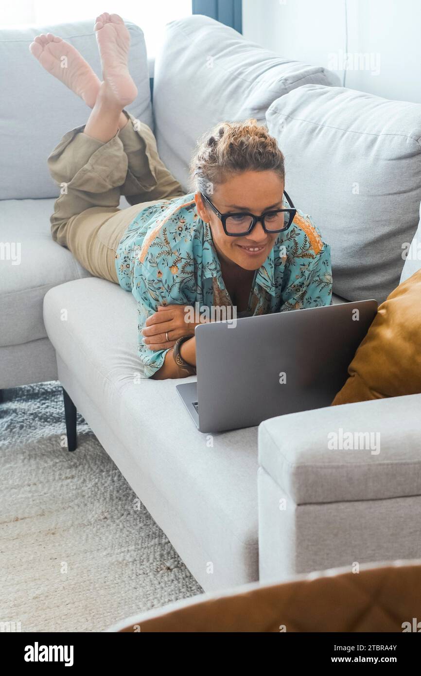 Intelligentes Arbeiten und Surfen im Internet. Eine Frau zu Hause, die auf dem Sofa liegt und sich entspannen kann, und die Technik im Innenbereich mit drahtlosem Internetzugang nutzen. Moderner Lebensstil mit Laptop auf der Couch Stockfoto