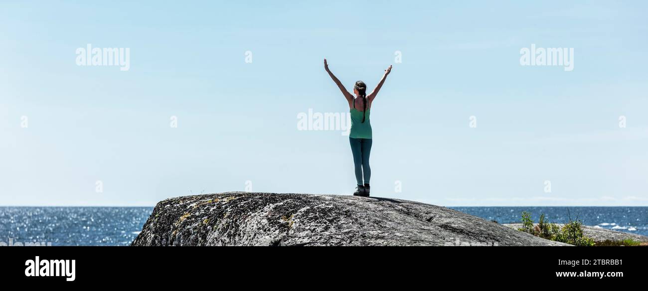 Frau mit ausgestreckten Armen auf einem felsigen Gipfel am Meer Stockfoto