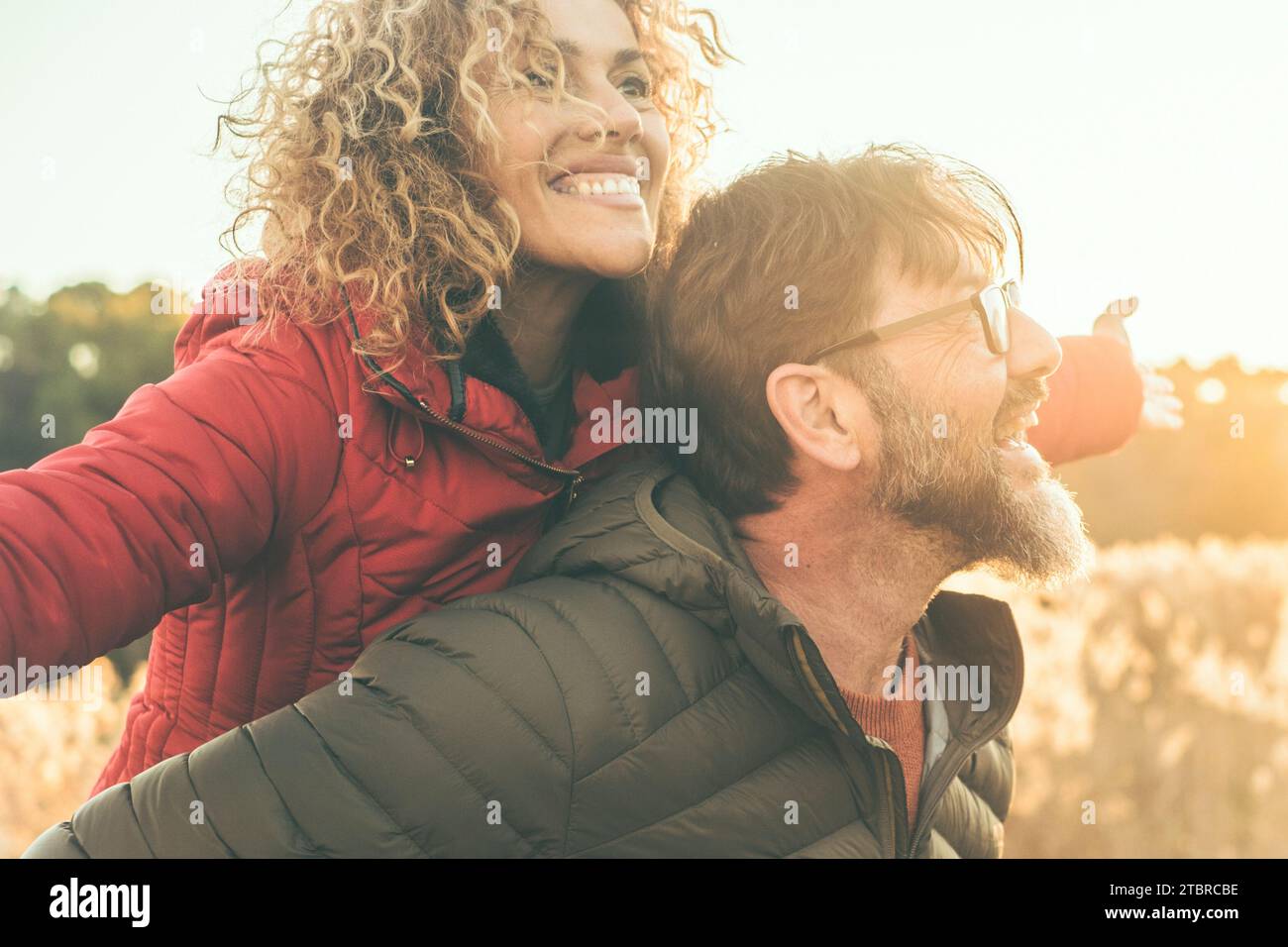 Glückliche Paare haben Spaß zusammen bei Freizeitaktivitäten im Freien im Naturgebiet während Sonnenuntergang und goldenen Stunden Licht. Der Mann trägt eine Frau auf dem Rücken. Menschen genießen das Leben und lachen. Winterherbsttag Stockfoto