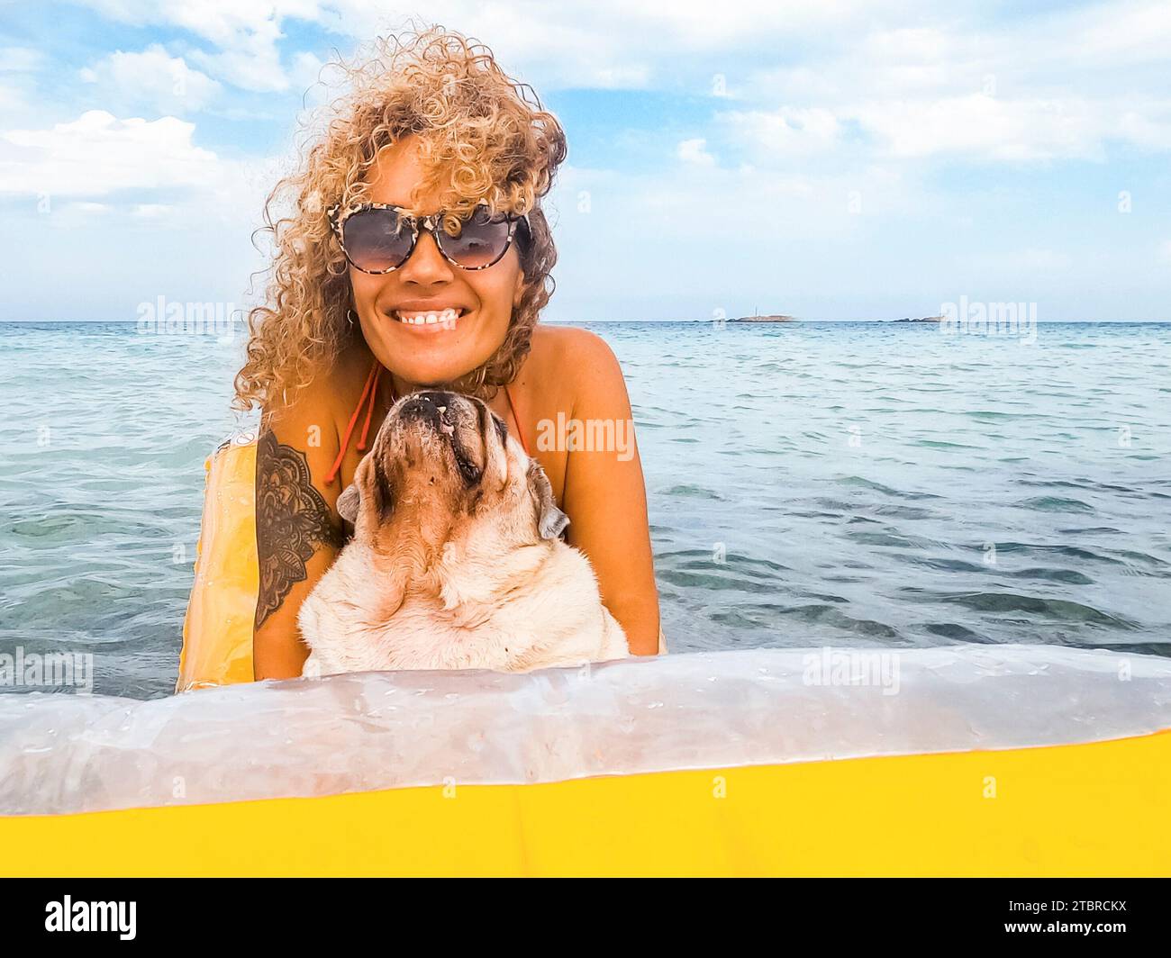 Glückliche Frau haben Spaß im Urlaub, spielen und entspannen auf einer gelben aufblasbaren Lilo mit ihrem Hund Mops darauf und schauen in der Kamera. Mensch und Tier Lebensstil. Urlaub mit Hund und Freizeit im Freien Stockfoto