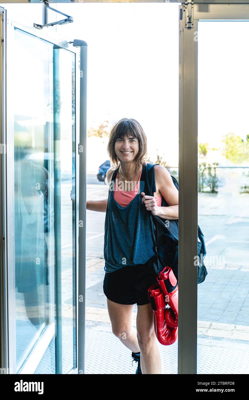 Vertikalfoto kaukasische Mittelerwachsene Frau mit Sportkleidung und -Ausrüstung, die lächelnd durch die Tür des Boxturniers hereinkommt. Sport und Erholung Stockfoto