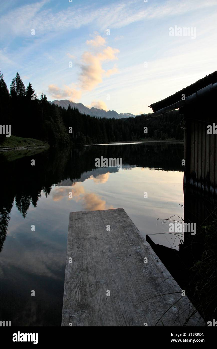 Bootshaus und Bootssteg bei Sonnenuntergang am Grubsee, Berge und Wald spiegeln sich im See, Blick auf die Wettersteinberge, Wetterstein, Krün, Oberbayern, Bayern, Deutschland Stockfoto