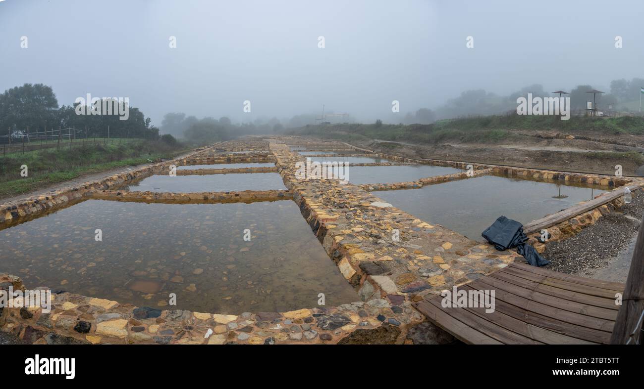 Panoramablick auf Salzverdunstungsteiche an einem nebeligen Tag, mit ruhigen Wasserflächen und rustikalen Barrieren. Stockfoto