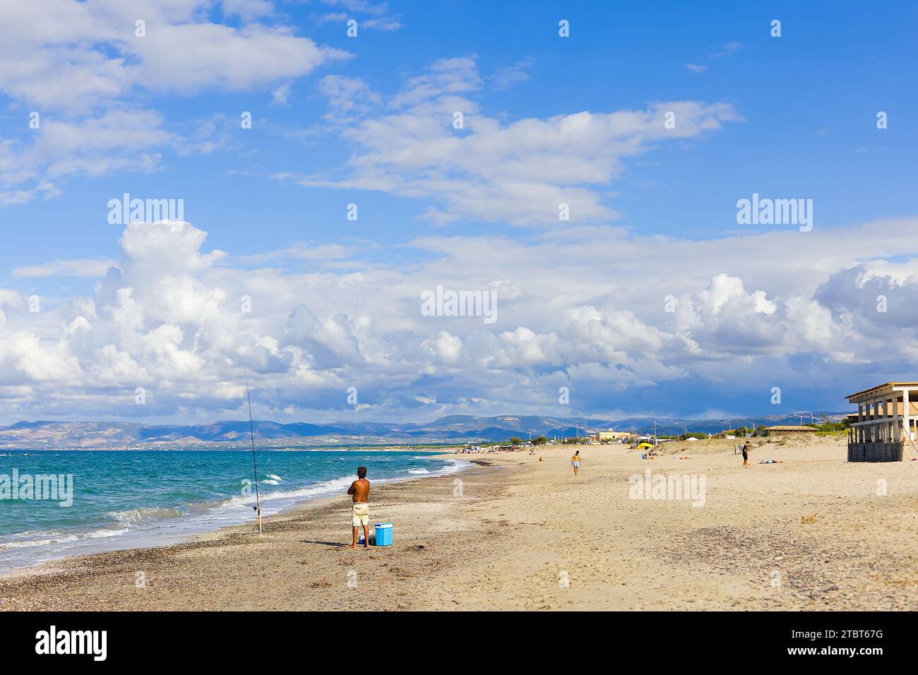 Am Strand Plata Mona, Sardinien, Italien, ist ein sardischer Fischer mit einer Rute abgebildet. Das Bild zeigt den Strand und die fernen Berge. Stockfoto