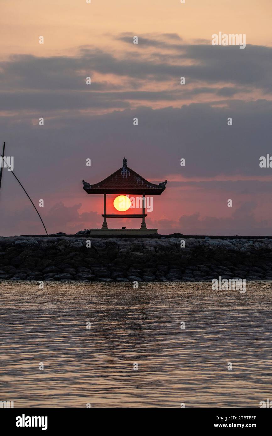 Sonnenaufgang am Sandstrand mit kleinen Tempeln im Wasser, Landschaft mit roter kreisförmiger Sonne am Strand von Sanur, Bali, Indonesien Stockfoto