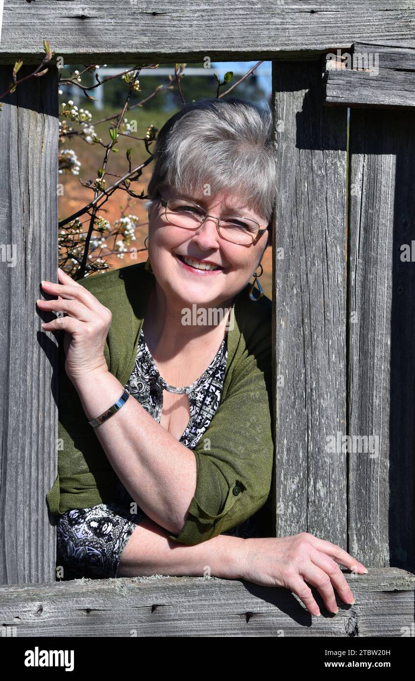 Die Frau nebenan schaut durch den kaputten Holzzaun. Sie trägt einen grünen Pullover und lächelt in der Frühlingssonne. Stockfoto