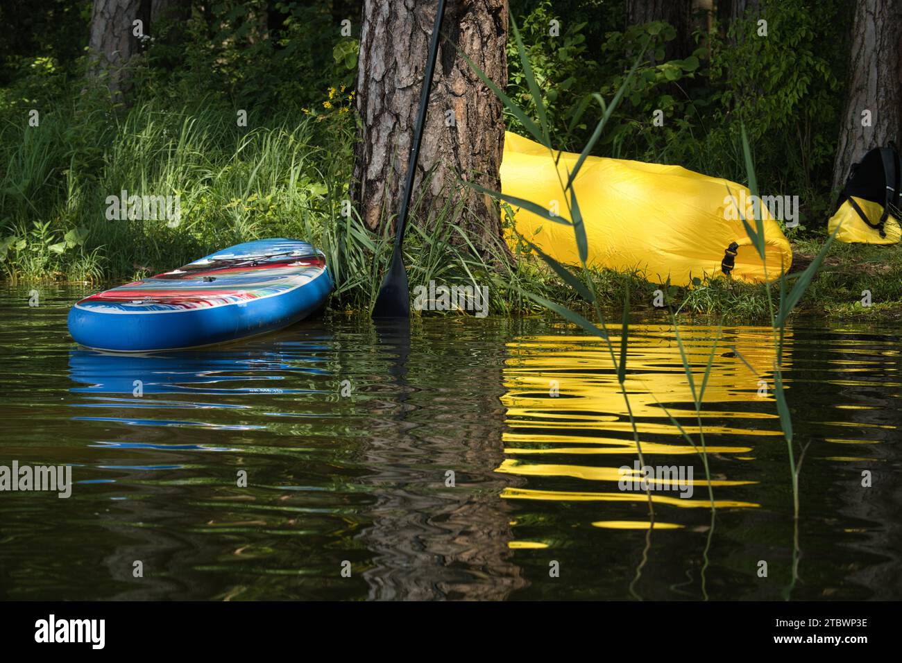 Stand up Paddleboard und gelbe aufblasbare Liege am Ufer des Sees im Schatten der Bäume und Reflexionen im ruhigen Wasser Stockfoto