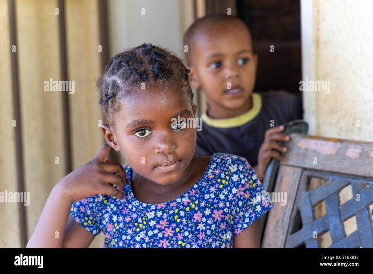 afrikanisches Mädchen mit Zöpfen und ihr Bruder spielt auf einer schmiedeeisernen Bank vor dem Haus Stockfoto