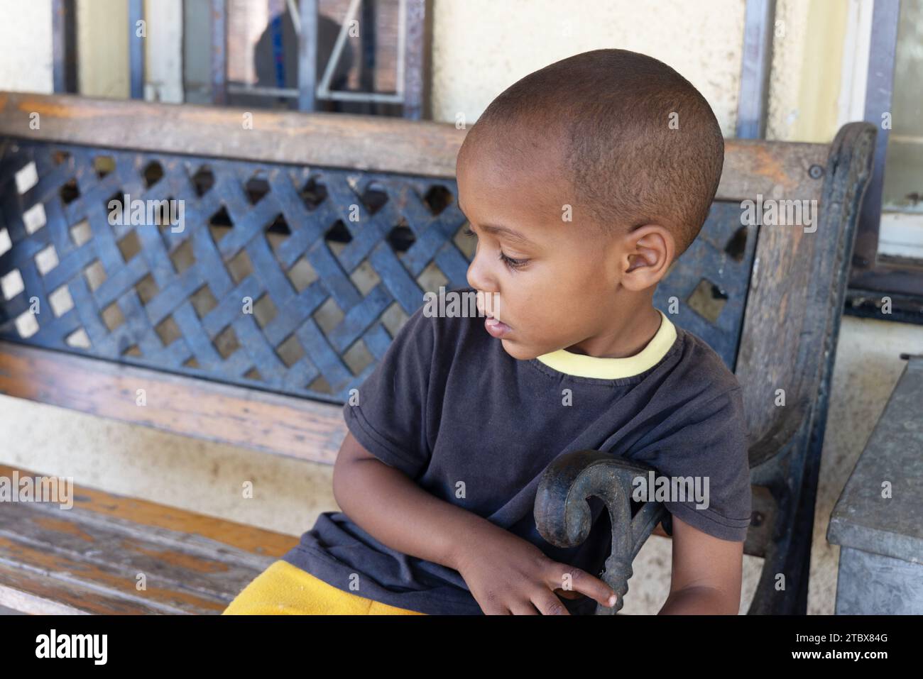 afrikanisches Kleinkind mit rasiertem Kopf, das auf einer schmiedeeisernen Bank vor dem Haus spielt Stockfoto