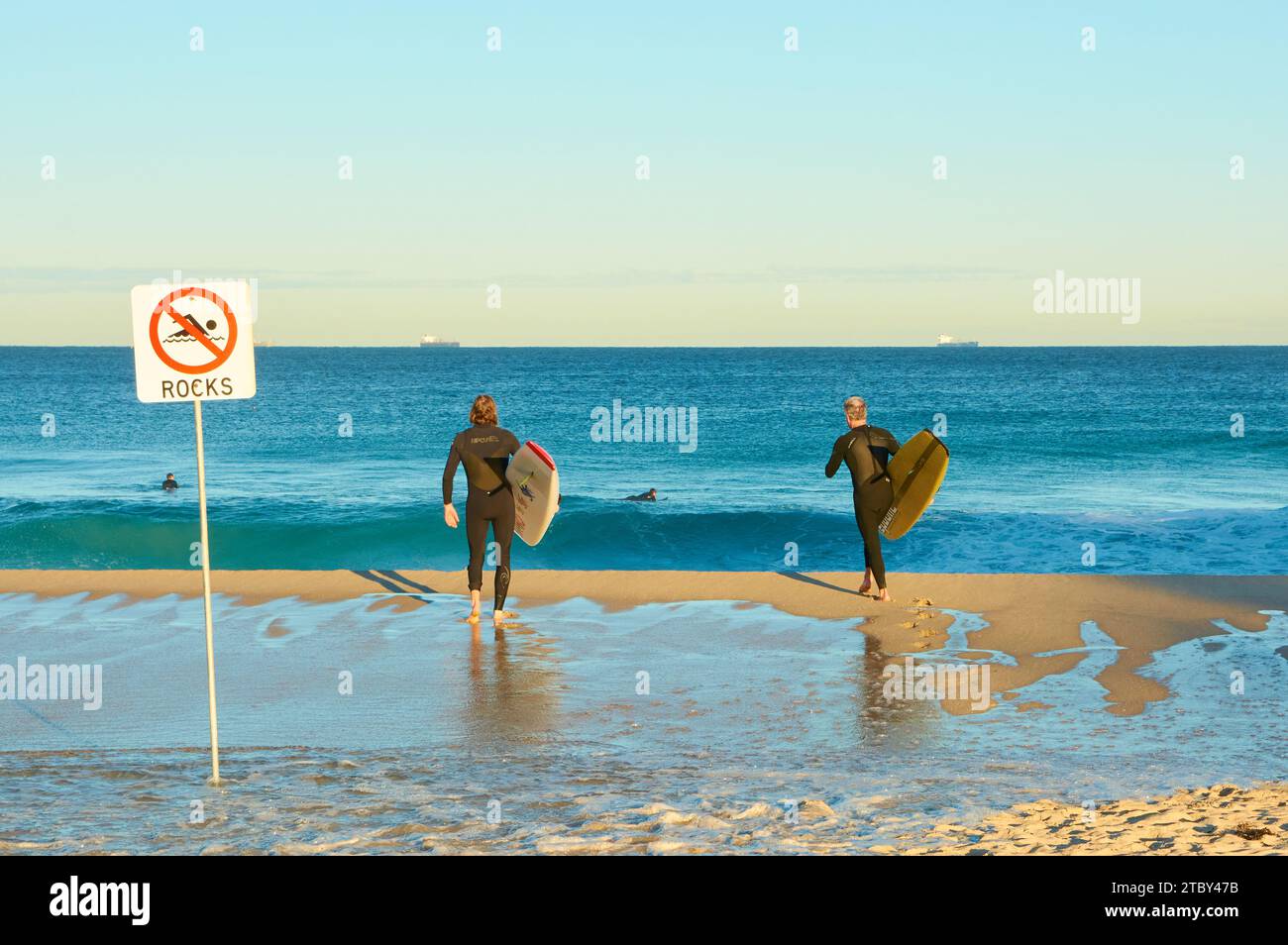 Zwei Männer, die mit ihren Brettern in den Ozean gehen, in der Nähe eines Schildes, das auf die Anwesenheit von Felsen hinweist, in Perth, Western Australia. Stockfoto