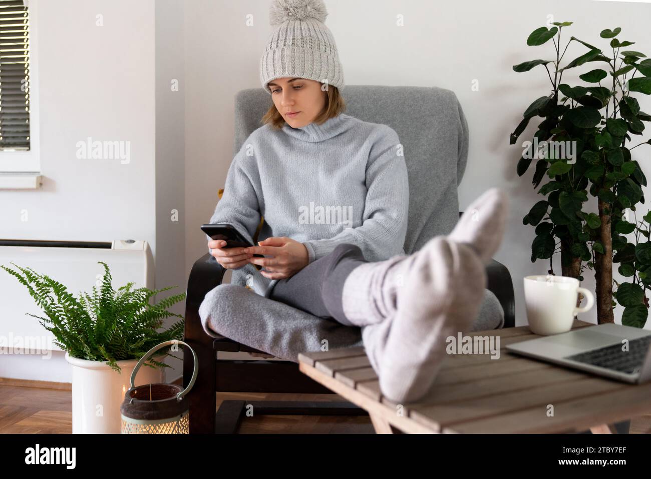 Eine Frau in warmer Kleidung mit Smartphone sitzt in der Nähe eines Heizstrahlers. Niedrige Temperatur im Haus. Energiekrise-Konzept. Stockfoto