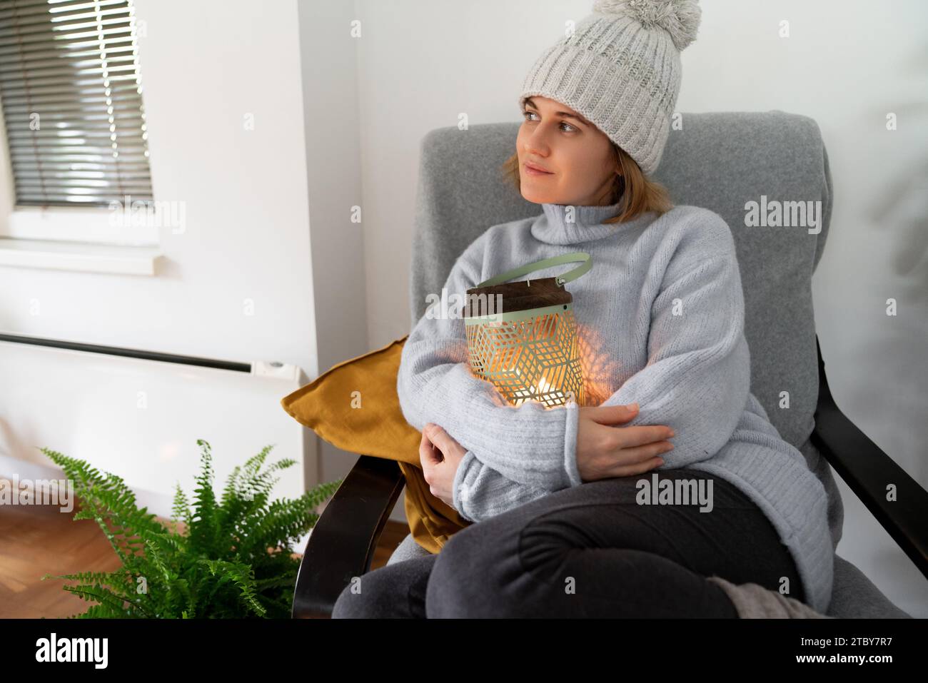 Eine Frau in warmen Kleidern sitzt in einem Sessel und umarmt eine Laterne mit Kerze. Niedrige Temperatur im Haus. Energiekrise-Konzept. Stockfoto