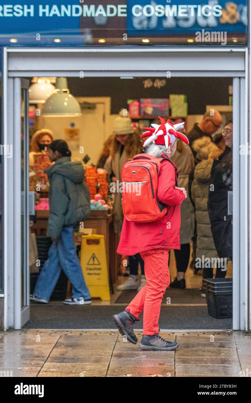 Mann mit roter festlicher Neuheit Jester Hut und Kleidung in Preston, Großbritannien. Dezember 2023. Weihnachtsläden, Shopper, Einkaufen an einem nassen Tag in der Fishergate High Street. Kredit ; MediaWorldImages/AlamyLiveNews Stockfoto