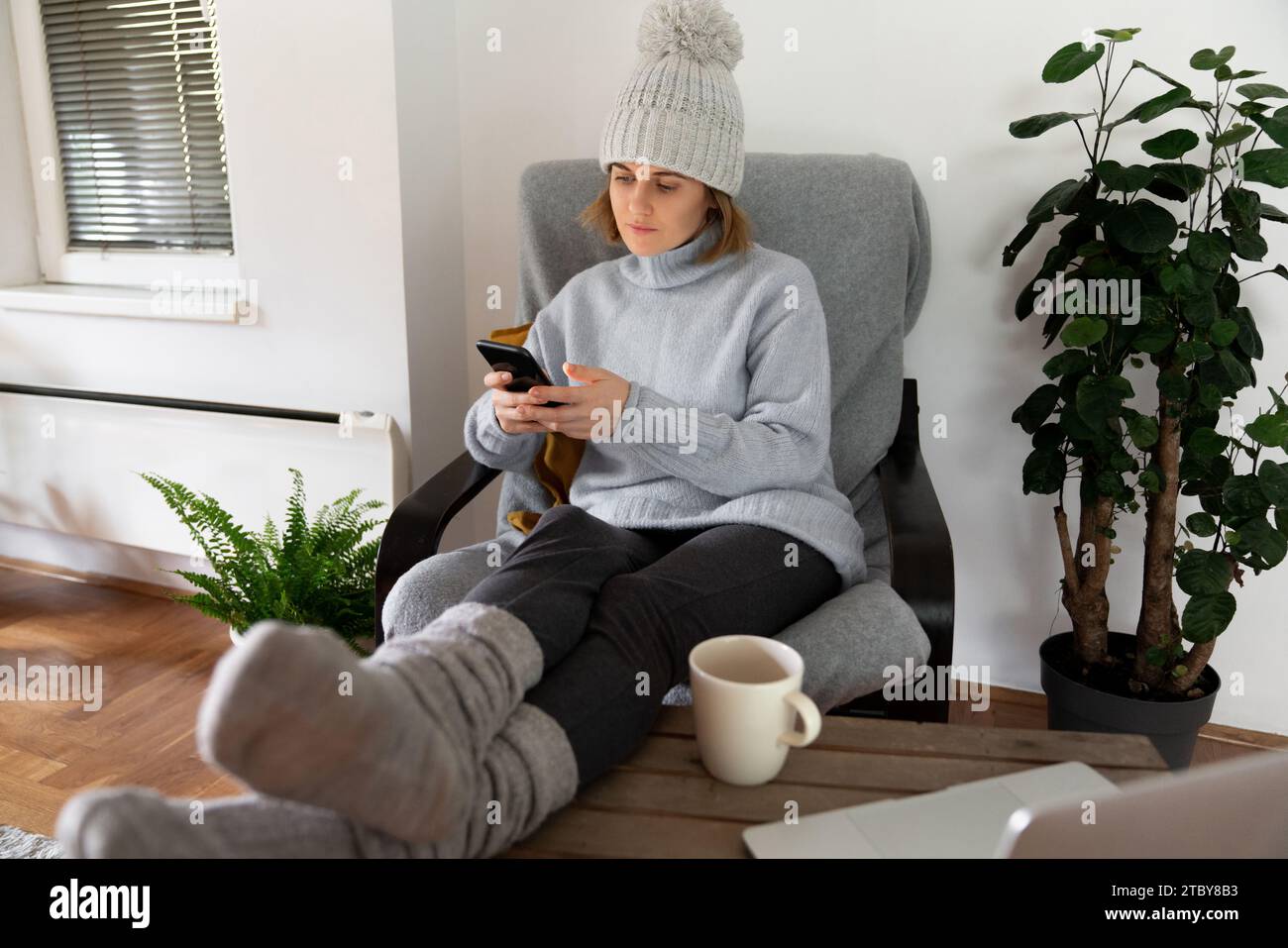 Eine Frau in warmer Kleidung mit Smartphone sitzt in der Nähe eines Heizstrahlers. Niedrige Temperatur im Haus. Energiekrise-Konzept. Stockfoto