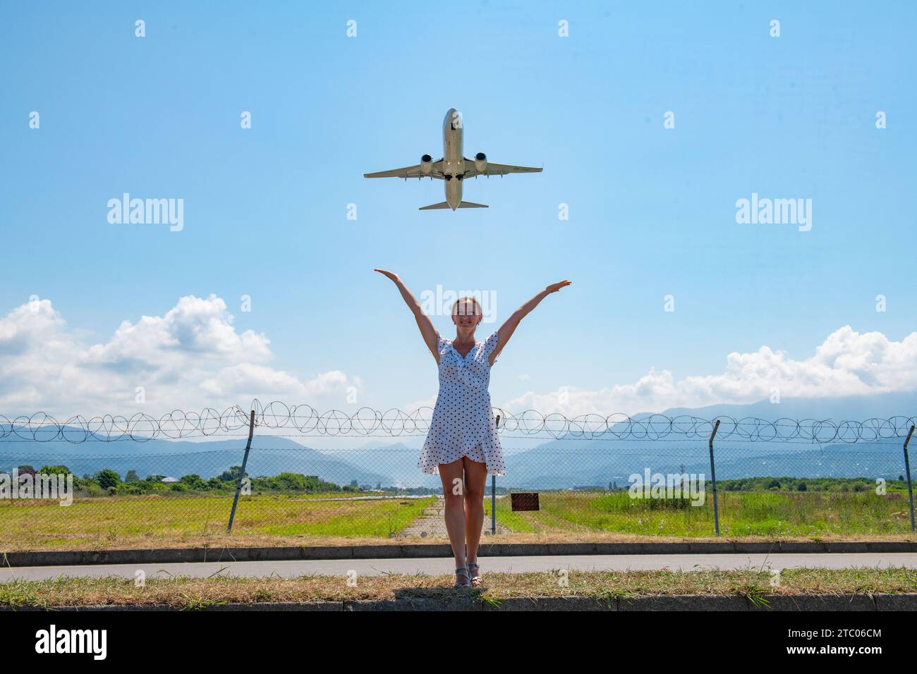Mädchen in weißem Kleid, das auf der Straße steht und ihre Hände am Himmel in Richtung Flugzeug streckt Stockfoto