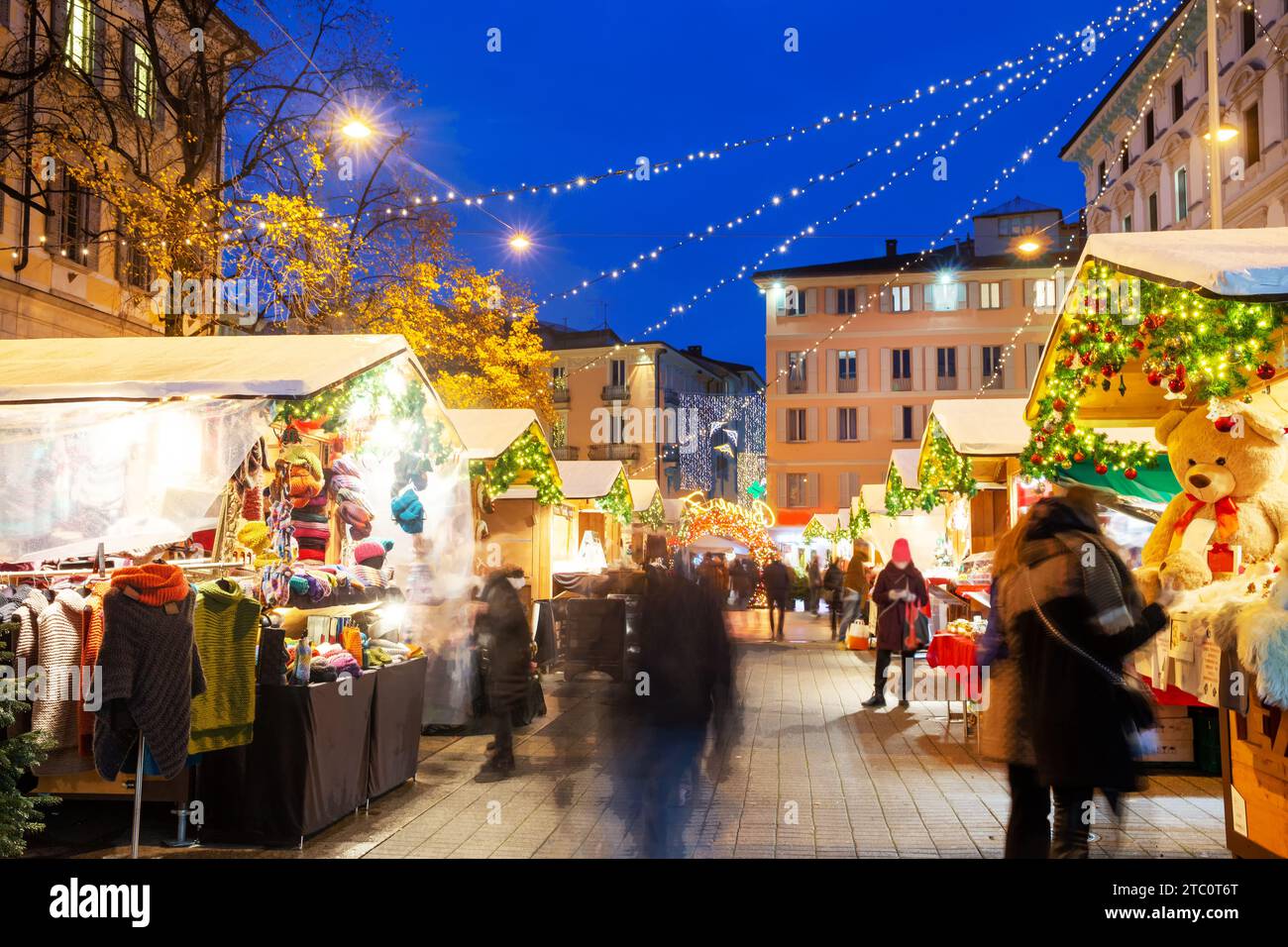 Weihnachtsmarkt mit Weihnachtsdekoration in Lugano neben dem Stadtplatz mit Rathaus. Verschwommene Bewegung Menschen in blauer Stunde - Dämmerung. Stockfoto