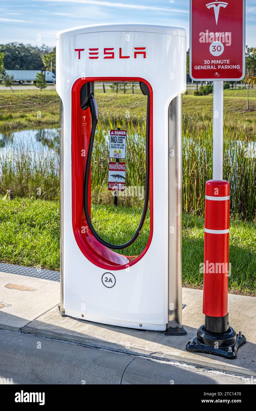 Tesla Supercharger EV-Station entlang der I-95 bei Buc-ees in Datona Beach, Florida, mit Alligator-Warnschild und interstate-Verkehr im Hintergrund. (USA) Stockfoto