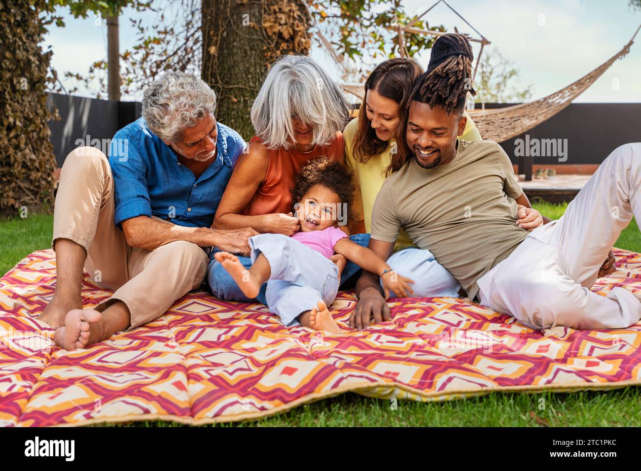 Die interrassische Familie mit mehreren Generationen teilt Lachen und Liebe auf einer farbenfrohen Picknickdecke und genießt einen fröhlichen Moment im Freien. Stockfoto