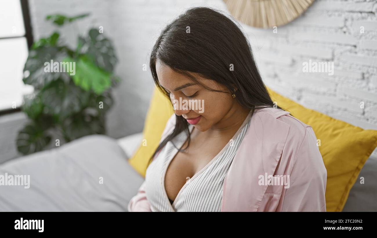 Eine junge schwangere Frau, die sich der Mutterschaft anschließt, sitzt entspannt auf dem Bett, blickt ernsthaft auf ihren Bauch in ihrem gemütlichen Schlafzimmer und denkt über den Morgen nach Stockfoto