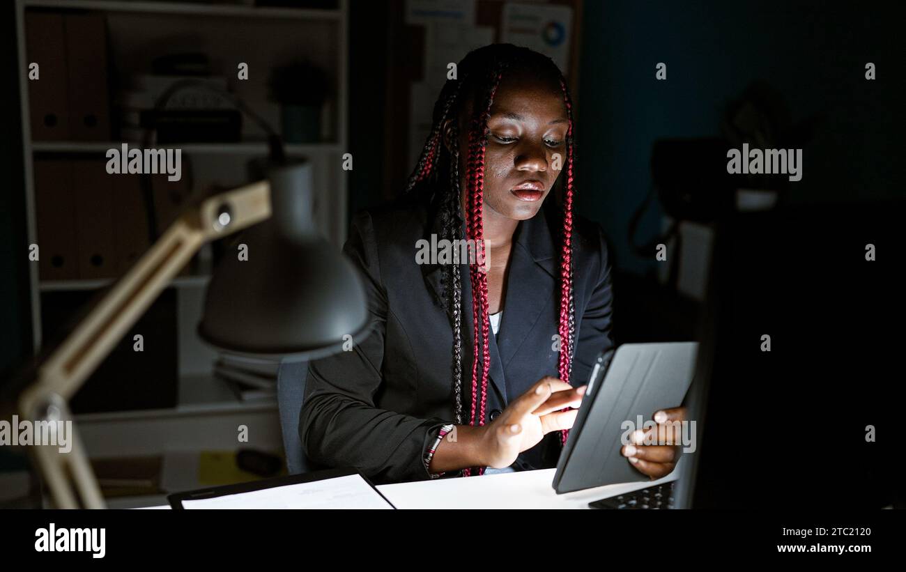 Fokussierte afroamerikanische Arbeitnehmerin im Büro, Monitore, Flechte und dunkle Jacke an, flinke Finger über Touchpad und Computer, beispielhaft el Stockfoto