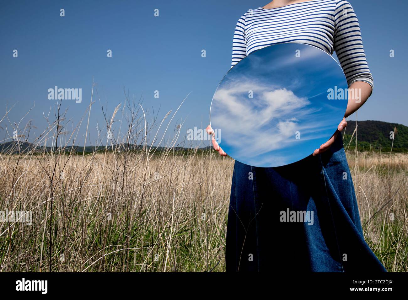 Surrealismus mit einer Frau, die einen Spiegel hält und ihr Gesicht auf dem Feld mit einem transparenten Hintergrund hinter dem Spiegel bedeckt Stockfoto