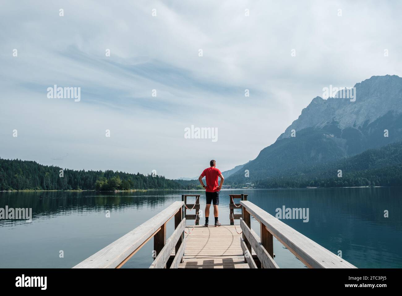 Blick vom Ufer des Eibsees in Deutschland, mit einem Wanderer in einem roten Hemd, der auf einer Fußgängerbrücke steht und in die Ferne blickt. Stockfoto