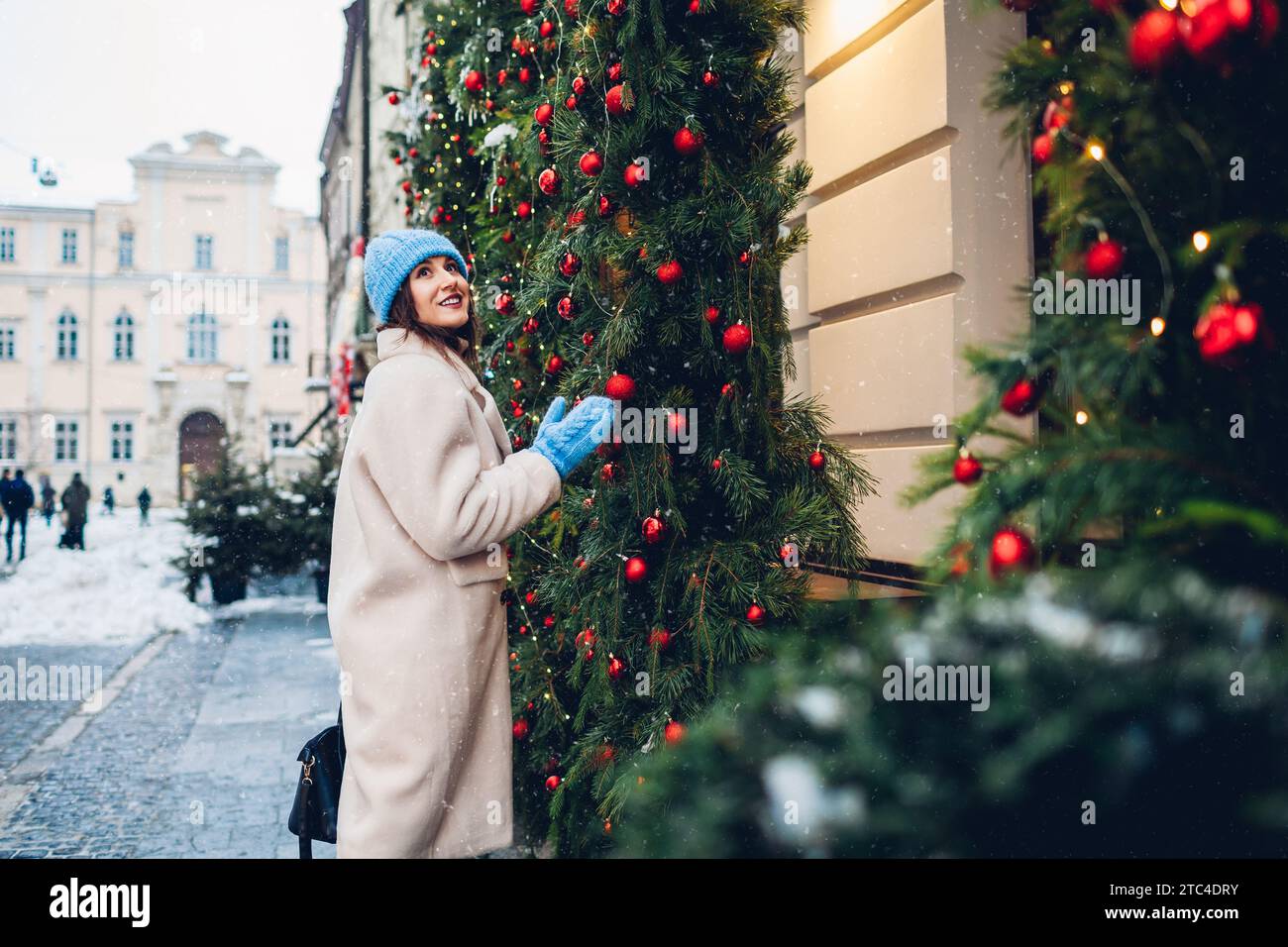 Weihnachtsstimmung. Glückliche Frau, die auf der Stadtstraße an Restaurants vorbeiläuft, die mit Tannengirlande und Ornamenten für Neujahr unter fallendem Schnee dekoriert sind Stockfoto