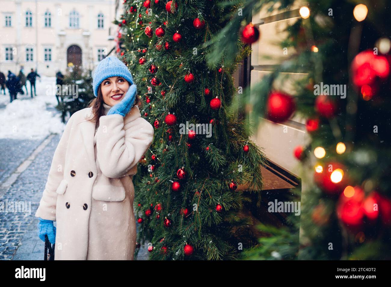 Weihnachtliche Atmosphäre. Lächelnde Frau, die auf der für Silvester geschmückten Stadtstraße unter fallendem Schnee spaziert und den Blick auf Girlanden, Lichter und Ornamente genießt Stockfoto