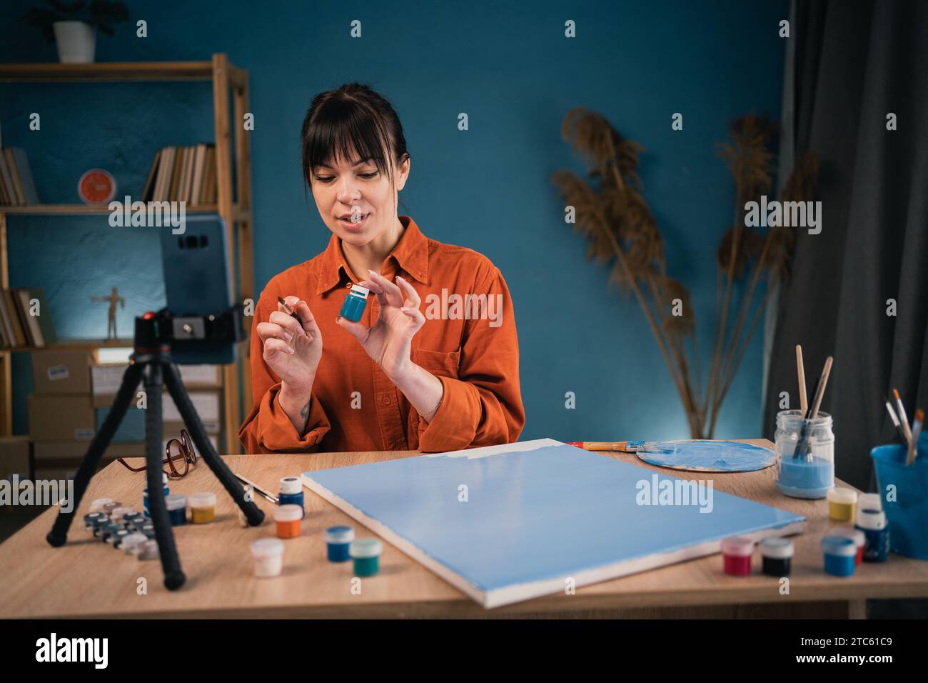 Schöne brünette Frau, die aus der Wohnung live streamt. Weibliche Künstlerin nimmt Video-Tutorial auf Smartphone über Kunstmalerei mit Stativ auf Stockfoto