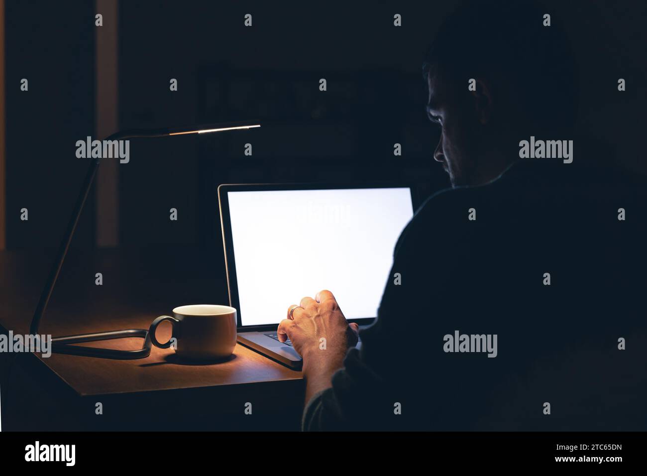 Ein Mann sitzt in einem dunklen Raum vor einem leeren Laptop-Bildschirm. Stockfoto