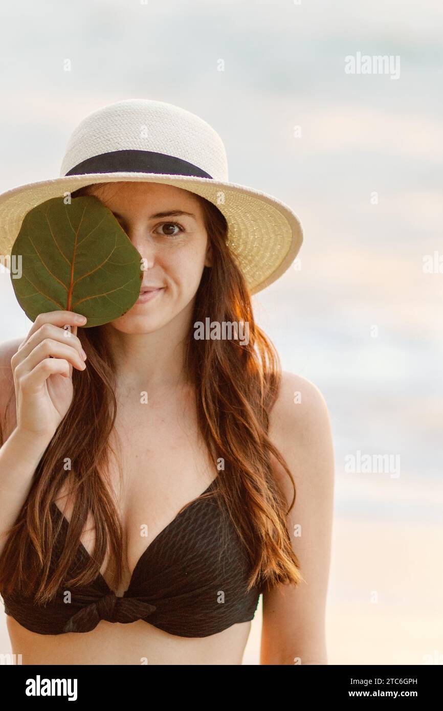 Entspannte Brünette in schwarzem Bikini und Sonnenhut, Blatt halten, bei Sonnenuntergang. Stockfoto